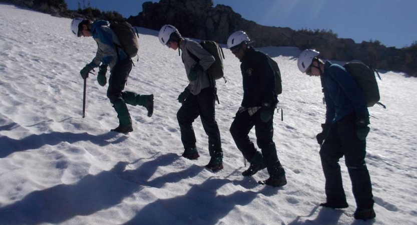 A group of outward bound students wearing mountaineering gear make their way up a snowy incline. 